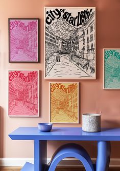 a blue table sitting in front of a pink wall with posters on the wall above it