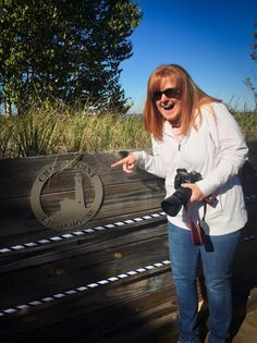 a woman standing next to a wooden bench pointing at the camera with her finger on it