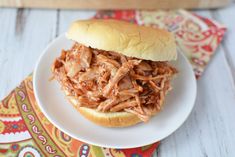 a pulled pork sandwich on a white plate with a colorful napkin and wooden cutting board in the background