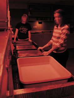 two people standing at a counter with trays in front of them and one person holding a spatula