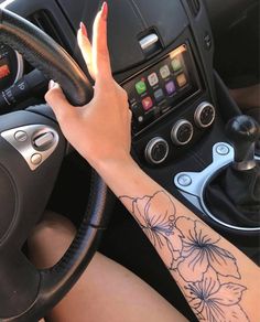 a woman driving a car with her hand on the steering wheel and flowers tattooed on her arm