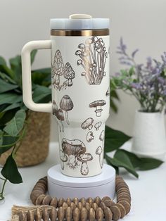 a white coffee cup with mushrooms on it sitting next to some plants and other items