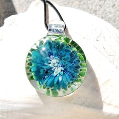 a blue and green flower is in a glass ball on a black cord necklace, sitting on a white rock