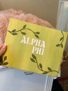 a person holding up a yellow book with the word alpha phi on it and a pink teddy bear