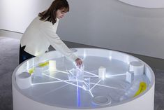 a woman standing over a glass table with various objects on it's surface and lights in the middle