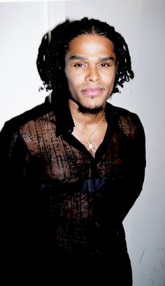 a man with curly hair standing in front of a white wall wearing a black shirt