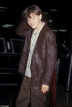 a young man standing in front of a car wearing a brown leather jacket and tan pants