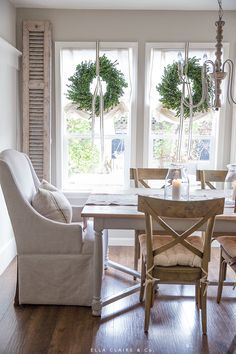 a dining room table with chairs and two wreaths hanging from the windows above it