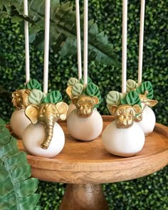 three white candles with gold decorations on top of a wooden stand in front of green foliage