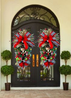 two christmas wreaths on the front door of a house with potted trees and bushes