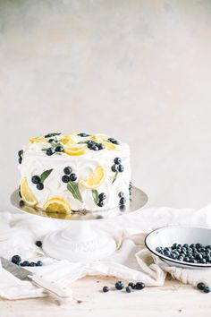 a white cake with blueberries and lemons on it sitting next to a bowl of blueberries