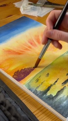 a person is holding a paintbrush and drawing on a piece of paper with an orange sky in the background