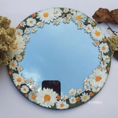 a mirror with flowers painted on it next to dried leaves and a cell phone in the foreground