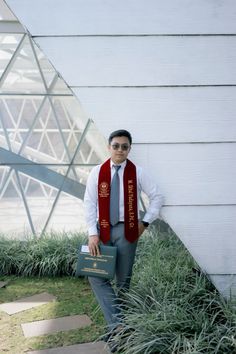 a man standing in front of a white building wearing a red scarf and grey pants