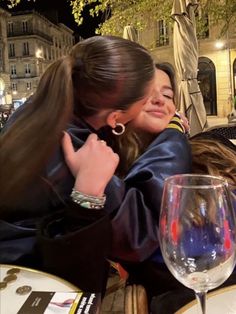 two young women hugging each other at a table with wine glasses on it and buildings in the background