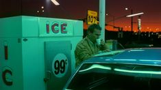 a man standing next to a blue car at a gas station with an ice machine