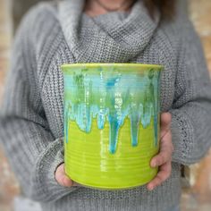 a woman holding a yellow and green cup with blue drips on the bottom, in front of a brick wall