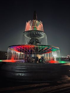 people are standing in front of a water fountain at night with colorful lights on it