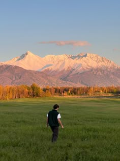 a man walking across a lush green field with mountains in the background on a sunny day