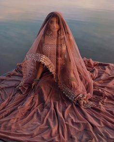 a woman sitting on top of a blanket next to a body of water wearing a veil