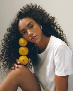 a woman with curly hair sitting on the ground holding three lemons