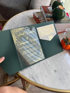 a person holding up a green and gold wedding card on top of a marble table