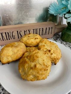 three biscuits on a white plate next to a vase with blue flowers and a sign that says baking is my therapy