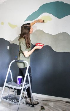 a woman is painting a mountain mural on the wall with a ladder and paint roller