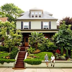 two people sitting on a wall in front of a house with plants and trees around it