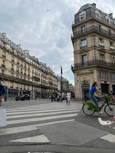 two people riding bicycles in the middle of a crosswalk with buildings on both sides