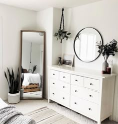 a bedroom with white furniture and plants on the dresser next to a large round mirror