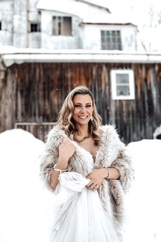 a woman standing in the snow wearing a white dress and fur stole with her arms crossed