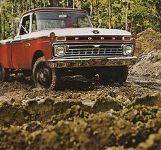 an old red and white truck is driving through the mud in front of some trees