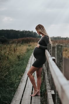 a pregnant woman standing on a wooden bridge in the middle of a field with tall grass