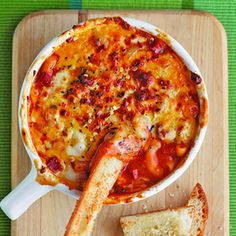 a wooden cutting board topped with bread and a bowl filled with cheese pizza dip next to a slice of garlic bread