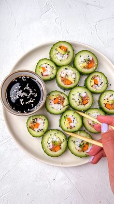 a plate with cucumber rolls and dipping sauce on it next to a hand holding chopsticks