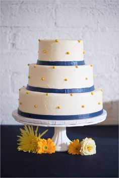 a three tiered white cake with blue and yellow decorations on the top, sitting on a table next to flowers