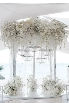 a white wedding setup with flowers and chandelier hanging from the ceiling in front of an ocean view