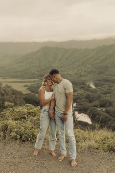 a man and woman standing next to each other on top of a hill with mountains in the background