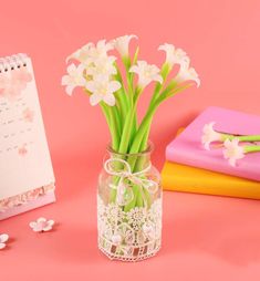 flowers in a vase next to a notepad and calendar on a pink table top