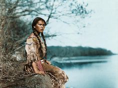 a native american woman sitting on a rock by the water