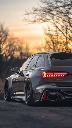 the rear end of a grey sports car parked in front of some trees at sunset