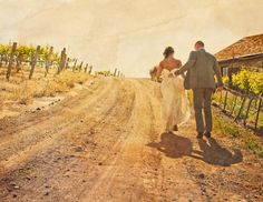 a bride and groom walking down a dirt road in front of a vineyard with yellow flowers