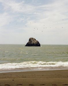 a rock sticking out of the ocean with birds flying over it in the distance on a cloudy day