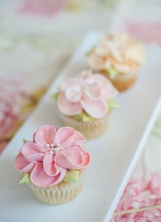 three cupcakes decorated with pink flowers on a white cake plate sitting on a floral tablecloth