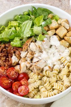 a white bowl filled with pasta salad and dressing next to sliced tomatoes, lettuce and croutons