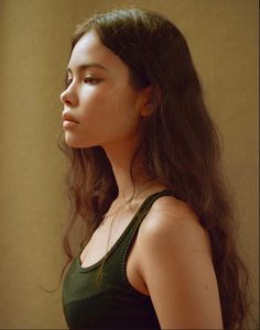 a woman with long dark hair standing in front of a beige wall and looking off to the side