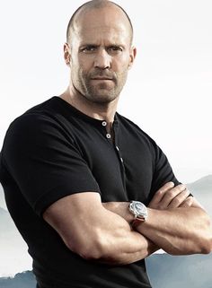 a bald man with his arms crossed standing in front of mountains and clouds, wearing a black polo shirt