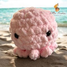 a pink crocheted stuffed animal sitting on top of a sandy beach next to the ocean