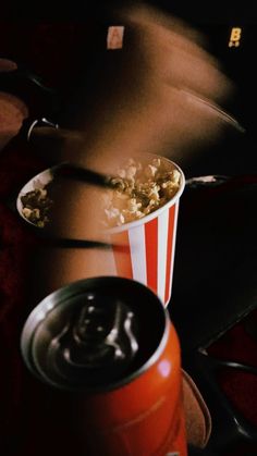 a red and white striped bucket full of popcorn next to a can of soda on the floor